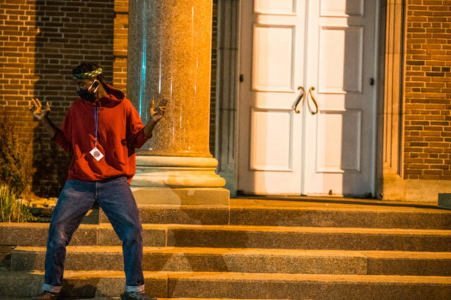 young black man in red sweatshirt and mask dances on the steps of a brick building.
