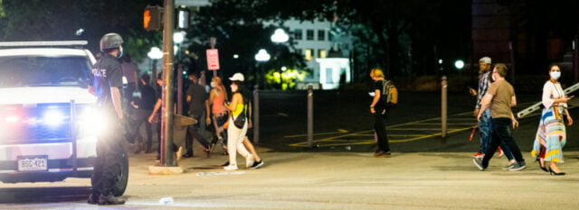 police officer stands in front of a parked police car with lights on. pedestrians in masks cross the street in a crosswalk