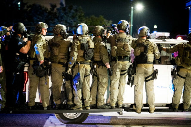 police officers and national guard in riot gear ride on the running boards of a denver police vehicle