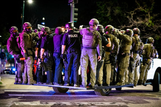 police officers and national guard in riot gear ride on the running boards on the side and back of a denver police vehicle