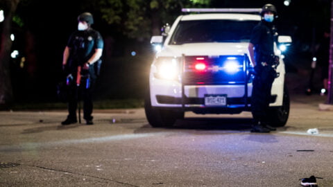 Parked police vehicle with lights on faces camera with two uniformed officers on either side of the police car.