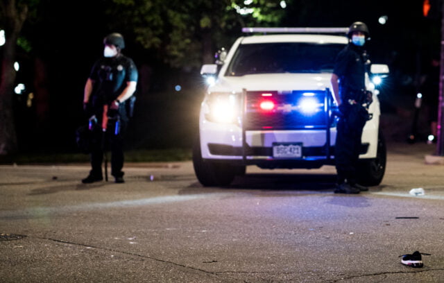 Parked police vehicle with lights on faces camera with two uniformed officers on either side of the police car.