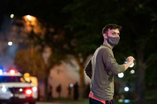pedestrian in a face mask walking in front of a parked police car with lights on. Background is lit by street light and smoke and protesters can be seen