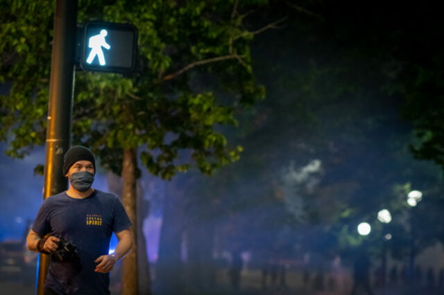 Night time photographer with hat and mask walking away from park/protest positioned under a "walk" street sign with smoke and pedestrians in the background