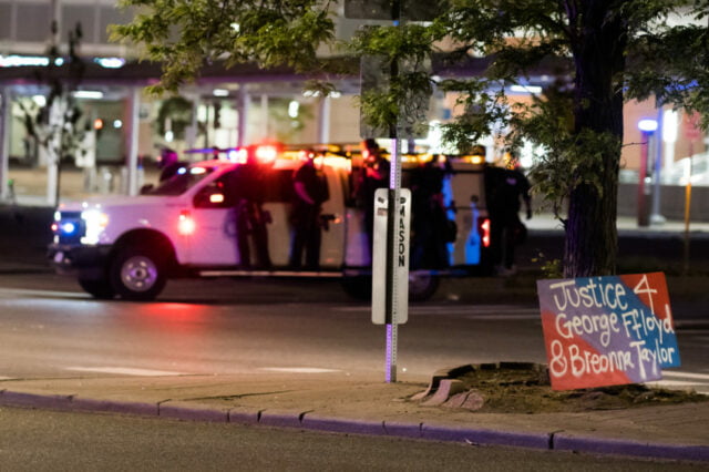 Corner of Colfax and Lincoln, police officers ride on the running boards of the police vehicle. Protest sign in foreground reads "Justice 4 George Floyd & Breonna Taylor"