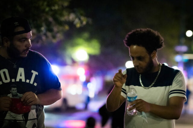 Two men of color engaged in conversation as they walk away from protest. with protest in background.
