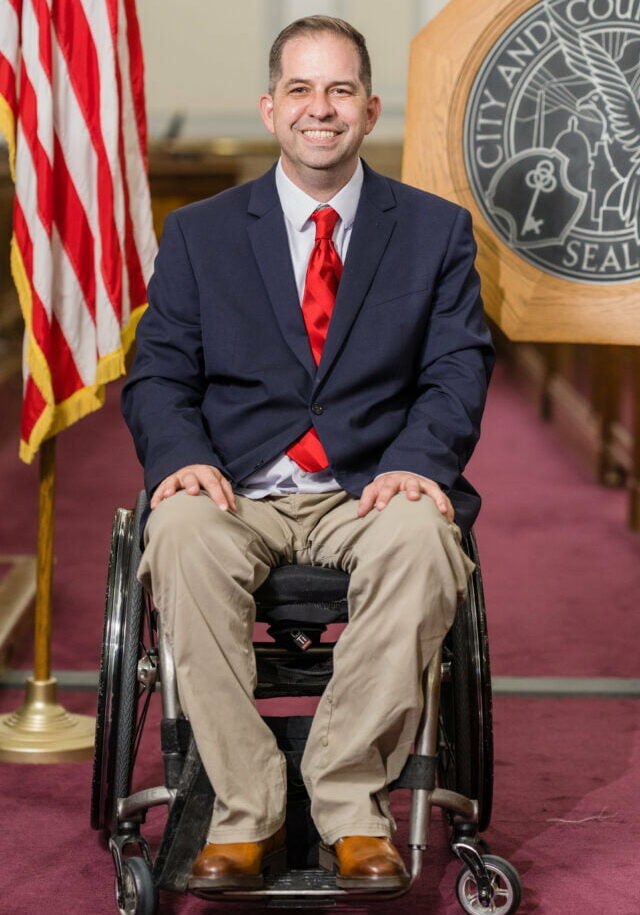 Photo of Chris Hinds in the City Council Chambers. He is wearing a blue shirt, dark blue coat, and red tie. He is sitting in a wheelchair. A US flag is behind his right shoulder, and the Denver seal is behind his left shoulder.